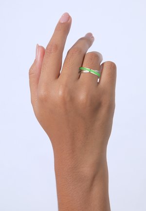 Sølv- og grøn crossover-ring på en hånd, der præsenterer en glat tekstur og et minimalistisk design; den grønne ring fremhæves mod sølvet.