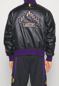 Chaqueta bomber de satén negra que presenta el texto bordado "LOS ANGELES LAKERS" en púrpura y dorado. Cuello y puños acanalados con rayas moradas.