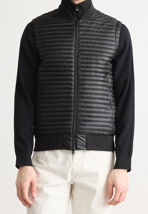 Gilet nero trapuntato con zip indossato sopra una camicia nera a maniche lunghe con pantaloni bianchi, mostrato dal collo ai fianchi.