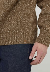 Pull en maille marron avec un motif texturé et des poignets côtelés. Présente une petite étiquette en cuir marquée "BZB" au bas. Jean foncé visible en dessous.