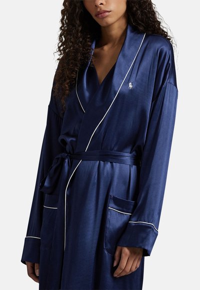 Vrouw draagt een marineblauwe satijnen badjas met witte biezen, gestrikt in de taille, met een klein geborduurd paardenlogo op de borst.