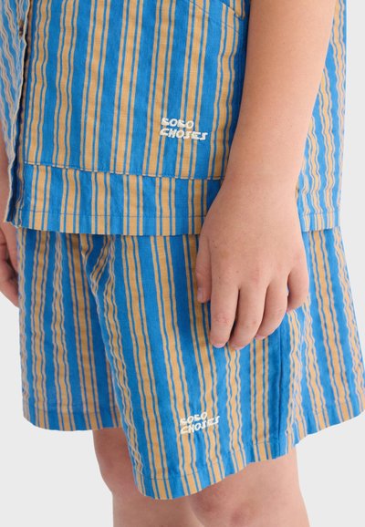 Shorts et chemise rayés bleu et jaune avec le texte brodé blanc "SOLO CHOICES", gros plan sur la main d'un enfant reposant à côté.