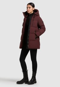 Bordó puffermellény, kapucnival, cipzárral és oldalsó zsebekkel. Simás textúra, steppelt kialakítás fekete részletekkel. Fekete leggings és csizmák.