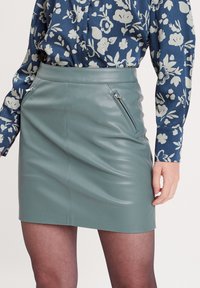 Mini-jupe en cuir vert avec une texture lisse, dotée d'une poche zippée sur le côté et de coutures minimalistes, associée à une chemise bleue à motifs.