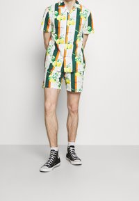 Conjunto de camisa de manga corta con botones y pantalones cortos, con un patrón de limones y flores sobre un fondo blanco con rayas verticales verdes y naranjas.