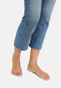 Pantalones capri de mezclilla en azul medio, con el dobladillo ligeramente deshilachado, usados con los pies descalzos. La tela muestra la textura típica de la mezclilla y un desgaste cerca de las rodillas.