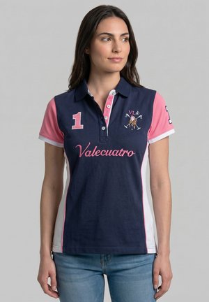 Mujer con un polo azul marino, rosa y blanco con "Valecuatro" y el número 1, de pie sobre un fondo gris liso.