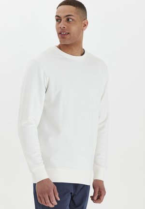 Felpa bianca a collo rotondo in cotone, con maniche lunghe e polsini a costine. Indossata con pantaloni blu navy. Design semplice, senza motivi.