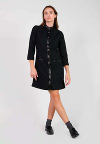 Vestido negro texturizado con cuello tipo camisa, botones en el frente y dos bolsillos laterales. Presenta mangas de tres cuartos y un acabado negro suave.