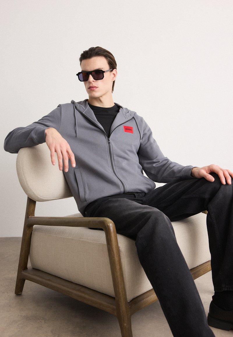 Siva zip-up dukserica s crvenim logotipom, crna majica, crne traperice. Model nosi tamne naočale dok sjedi na svijetloj stolici.