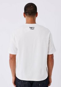 Camiseta blanca de manga corta con un logo negro de "Project x Paris" en la parte trasera del cuello. Fabricada con tejido suave y con un corte relajado.