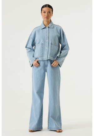 Vrouw staande, gekleed in een lichtblauwe denim jas met fijne strepen en bijpassende wijde jeans, met handen in de zakken tegen een witte achtergrond.