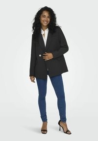 Blazer noir texturé avec un design à double boutonnage et une broche en argent, associé à un jean skinny bleu et des escarpins noirs à bout ouvert.