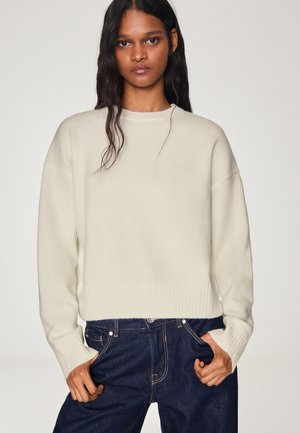 Femme aux longs cheveux foncés portant un pull en tricot crème et un jean bleu foncé, debout les mains dans les poches devant un fond uni.