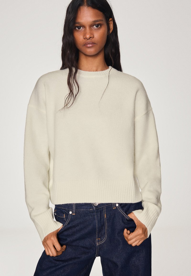 Femme aux longs cheveux foncés portant un pull en tricot crème et un jean bleu foncé, debout les mains dans les poches devant un fond uni.