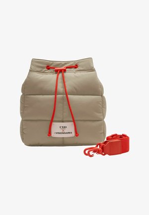 Sac seau beige matelassé avec fermeture à cordon rouge, écusson logo sur le devant, et bandoulière rouge amovible posée à côté.