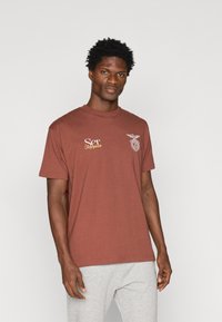 Καφέ βαμβακερό μπλουζάκι με κοντά μανίκια, με λευκό και χρυσό κείμενο και έμβλημα στο στήθος. Στρογγυλή λαιμόκοψη; λεία υφή.