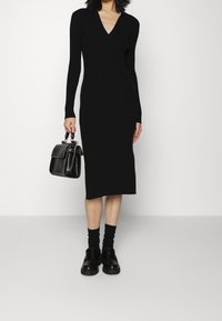Abito nero a costine con maniche lunghe e scollatura a V. Abbinato con scarpe nere chunky e una piccola borsa nera. Tessuto liscio.