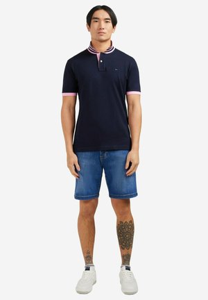 Polo bleu marine avec un col rayé rose et blanc, manches courtes avec des accents roses, associé à un short en denim bleu et des baskets blanches.