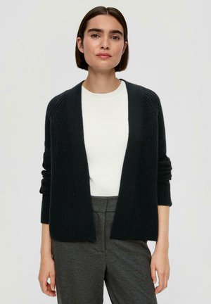 Schwarze Strickjacke mit offenem Vorderteil, geripptem Muster und langen Ärmeln über einem weißen Top. Kombiniert mit grauen, maßgeschneiderten Hosen.