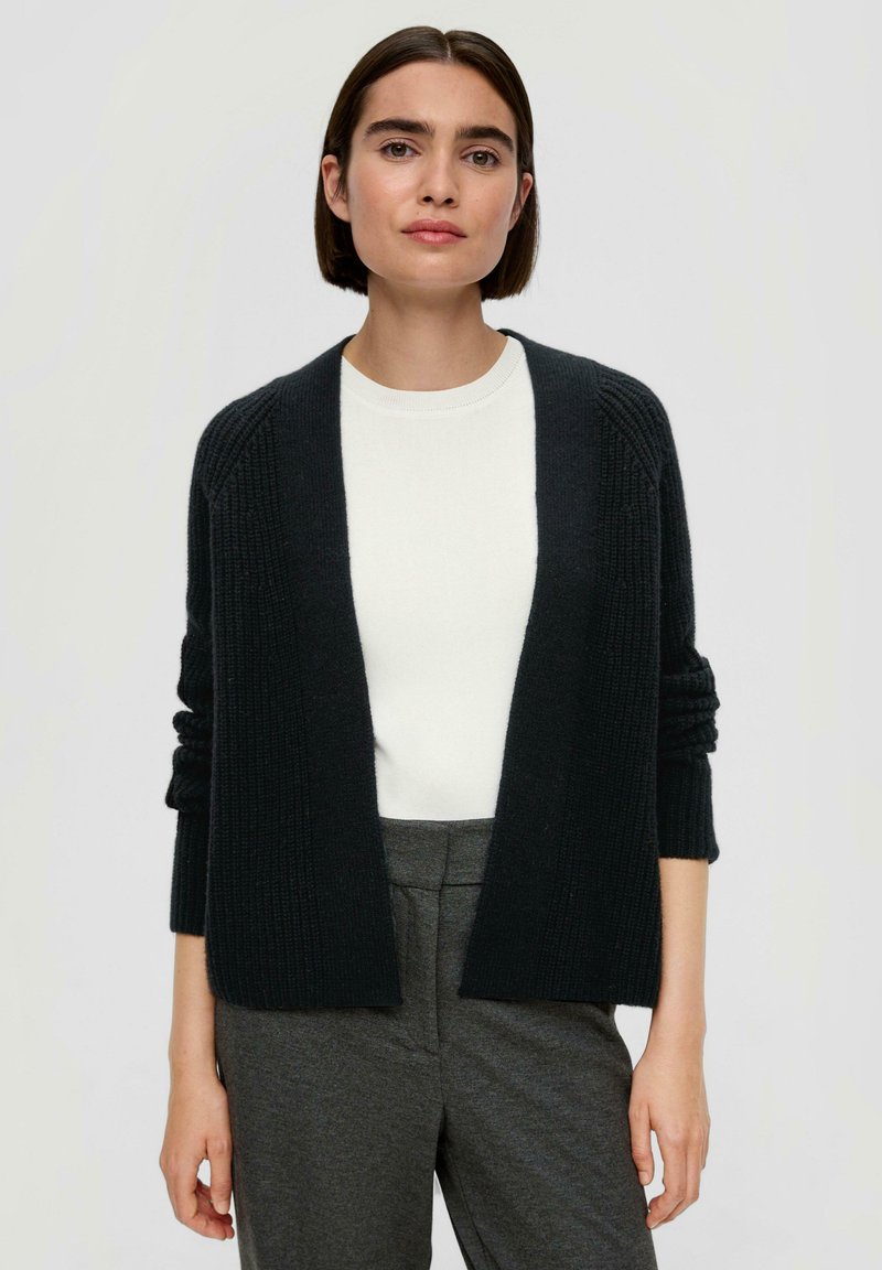 Schwarze Strickjacke mit offenem Vorderteil, geripptem Muster und langen Ärmeln über einem weißen Top. Kombiniert mit grauen, maßgeschneiderten Hosen.