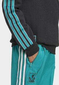 Felpa nera con strisce turchesi e polsini a costine abbinata a pantaloni turchesi con strisce bianche e patch del Liverpool Football Club.