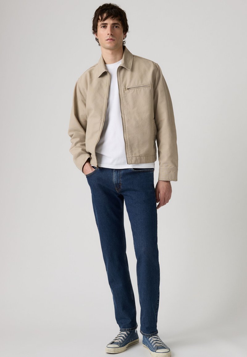 Giovane uomo in piedi con la mano in tasca che indossa una giacca beige, una t-shirt bianca, jeans blu e sneakers blu su sfondo semplice.