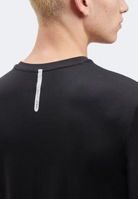 Κοντινό πλάνο ενός ατόμου με κοντά ξανθά μαλλιά που φοράει μαύρο αθλητικό πουκάμισο με υφή και κάθετη λευκή ετικέτα "Ted Baker Sport" στο πάνω μέρος της πλάτης.