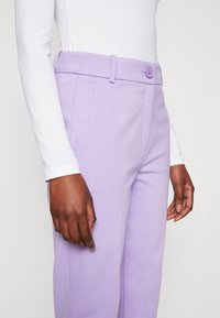 Lila broek met een gladde textuur, voorzien van een knoopsluiting en riemlussen, naast een witte top met lange mouwen.