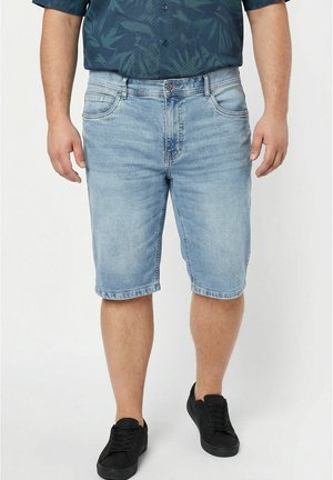 Mann trägt hellblaue Jeansshorts, ein dunkles gemustertes Hemd und schwarze Sneakers, steht vor weißem Hintergrund.
