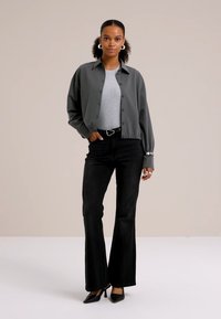 Chaqueta gris de botones sobre una camiseta gris, combinada con jeans de campana negros y tacones negros. Incluye herrajes plateados y tejido texturizado.