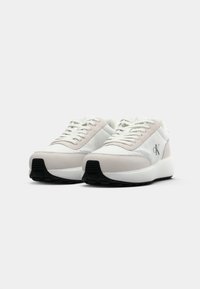 Sneaker bianchi e beige chiaro con suola nera, design con lacci e logo "cK" sul lato, presentati su uno sfondo bianco semplice.
