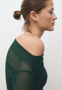 Bluză de culoare verde închis, cu umerii expuși, realizată dintr-un material transparent. Prezintă o textură netedă și un design ajustat, cu straturi vizibile în jurul umerilor.