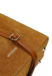 Hnědá semišová kabelka s koženým popruhem a kovovou sponou, s čtyřmi kovovými cvočky na spodním rohu a nápisem značky "wojas" ve stříbrných písmech.