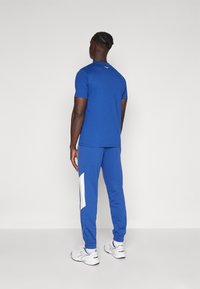 Blue athletic tracksuit with short-sleeve top and tapered pants. Pants have white side panel; fitted design, smooth fabric, and elastic cuffs.