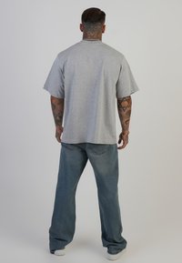 Pelēka īsām piedurknēm t-kreklu ar brīvu piegulšanu, apvienots ar gaiši zilu plata kāju džinsu un baltām kedām, skatīts no aizmugures.