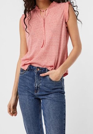 Kvinde iført ærmeløs pink top med flæset ærmegab og blå jeans med høj talje, hånd stukket i forlomme, mod ensfarvet baggrund.