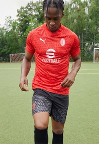 Maglia da calcio rossa con motivi triangolari e colletto a giro, abbinata a pantaloni corti neri con un motivo simile. Indossata con calzini neri.