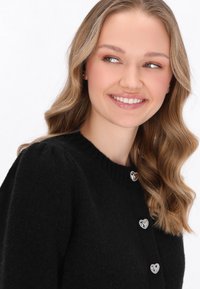 Černý pletený cardigan s kulatým výstřihem, který má stříbrné knoflíky ve tvaru srdce na přední straně. Měkká textura s těsným střihem.