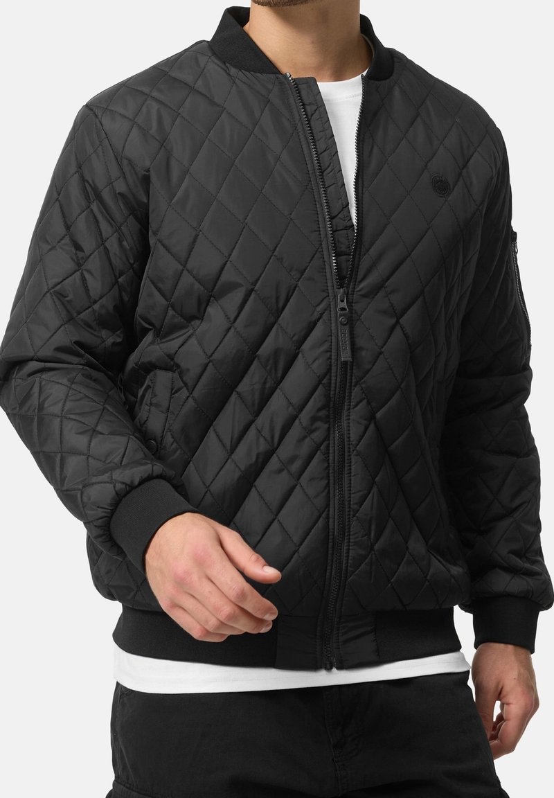 Homme portant une veste bomber matelassée noire avec une fermeture éclair frontale, des poignets et une taille côtelés, par-dessus une chemise blanche.