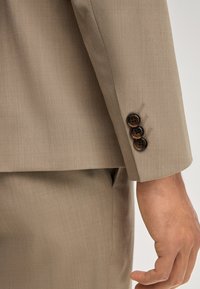 Beiger Anzugsärmel mit drei dunkelbraunen Knöpfen am Manschettenrand und passenden beigen Hosen, die einen Teil der Hand und des Handgelenks einer Person zeigen.