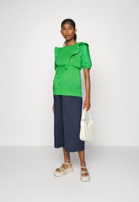 Top verde de manga curta com detalhes em babados, atado na cintura, combinado com culotes largos azul-marinho e sandálias de plataforma leves. Bolsa branca.