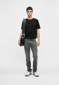 Jovem vestido com uma t-shirt preta, jeans cinzentos, ténis brancos com riscas pretas e a carregar uma grande bolsa de couro preta com uma alça grossa.