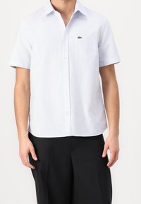 Vaaleansininen ja valkoinen raidallinen lyhythihainen paita, valmistettu kudotusta kankaasta, jossa on napitettava etuosa ja rintatasku, jossa logo.