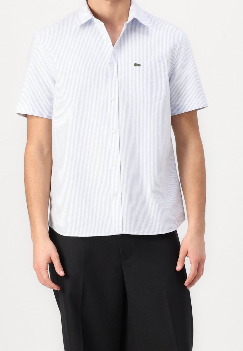 Vaaleansininen ja valkoinen raidallinen lyhythihainen paita, valmistettu kudotusta kankaasta, jossa on napitettava etuosa ja rintatasku, jossa logo.