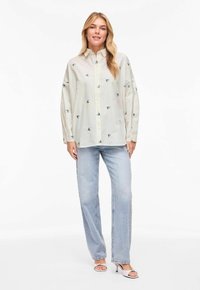 Femme portant une chemise blanche ample à boutons avec de petites impressions de fleurs bleues, un jean droit bleu clair et des talons blancs à bout ouvert, debout sur un fond blanc.