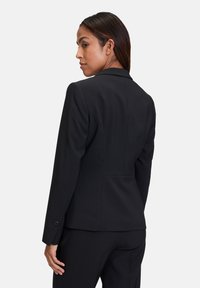 Černý vypasovaný blazer s úzkým střihem, klopovými límci a bočními rozparky. Obsahuje manžety na knoflíky a hladký povrch.