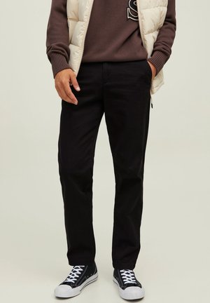 Hombre que lleva pantalones rectos negros, zapatillas negras y blancas, un suéter marrón y un chaleco acolchado crema, de pie con las manos en los bolsillos.