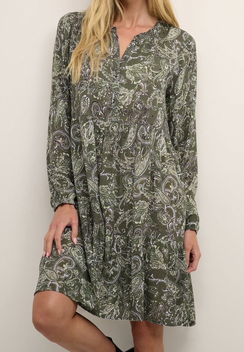 Vihreä paisley-kuosiset mekko, jossa on pitkät hihat, napitettava kaulus, rypistetty vyötärö ja teksturoitu kangas. Kuosi sisältää monimutkaisia lehtimuotoiluja.