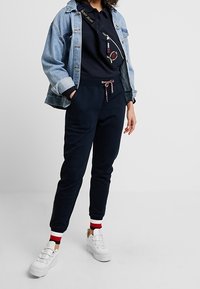 Pantaloni della tuta blu navy con coulisse, abbinati a una maglietta scura e una giacca di jeans strappata. Sneakers bianche con accenti rossi e bianchi completano il look.